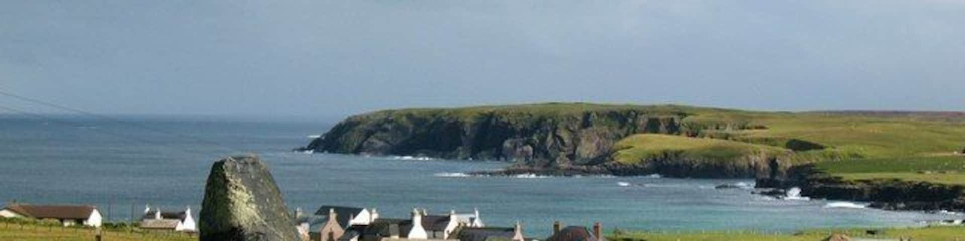 A coastal landscape with a prominent standing stone, grassy fields, and a small settlement with white-roofed buildings by the sea.