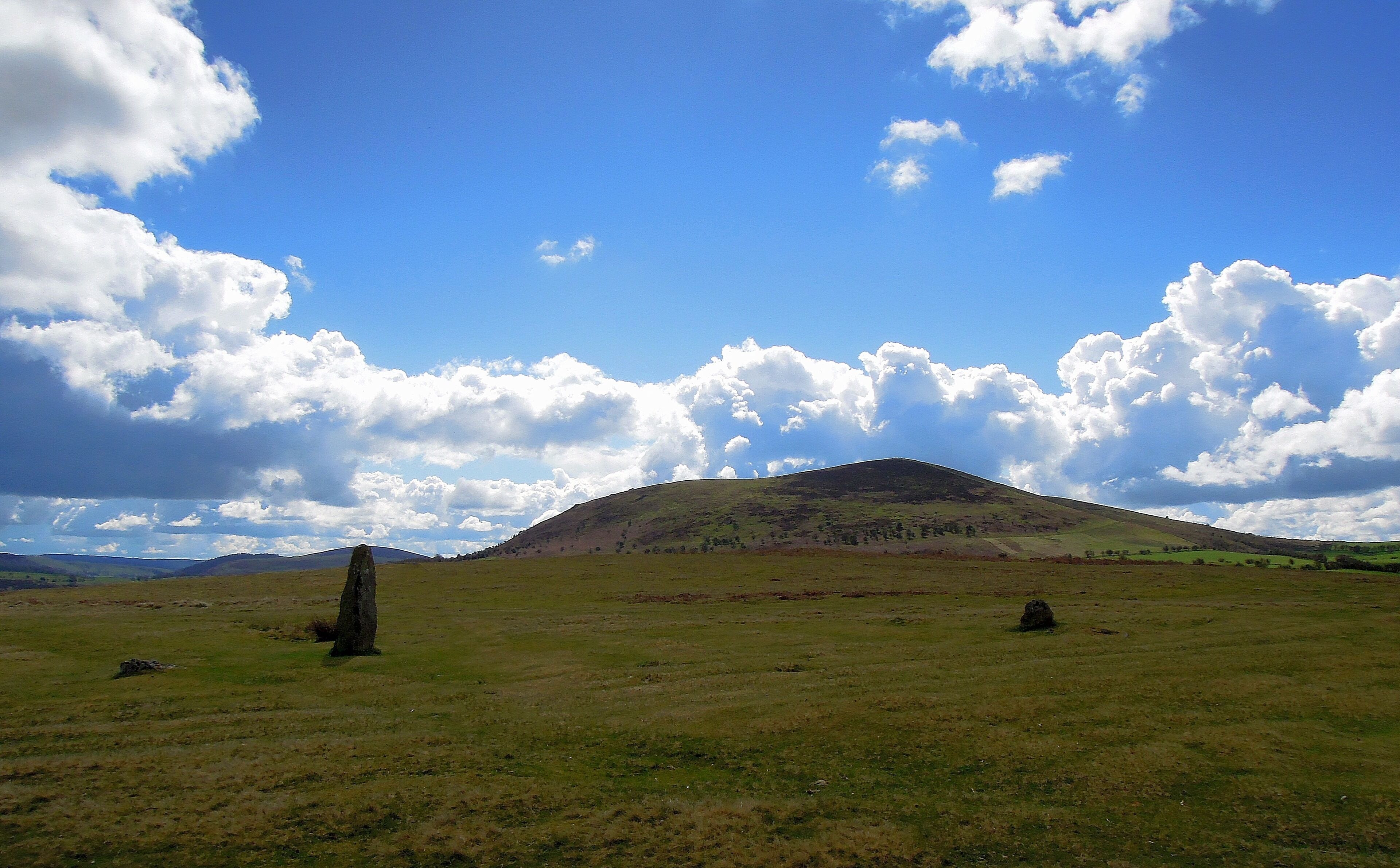 A grassy field with a prominent hill and a standing stone in the foreground.