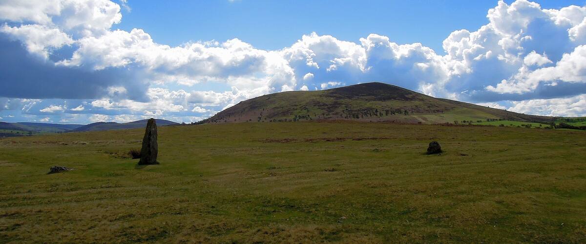 A grassy field with a prominent hill and a standing stone in the foreground.