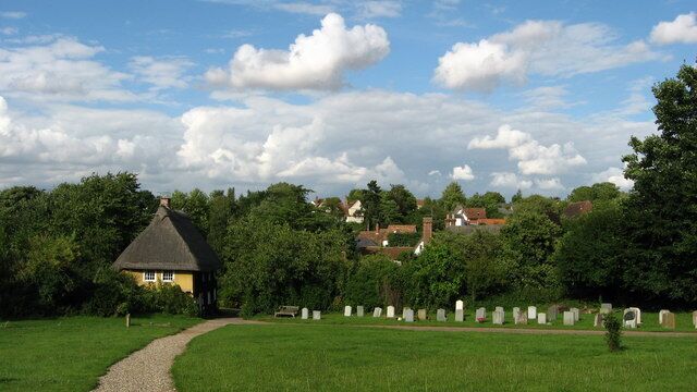 A rural cemetery with headstones, a thatched-roof house, and surrounding greenery.