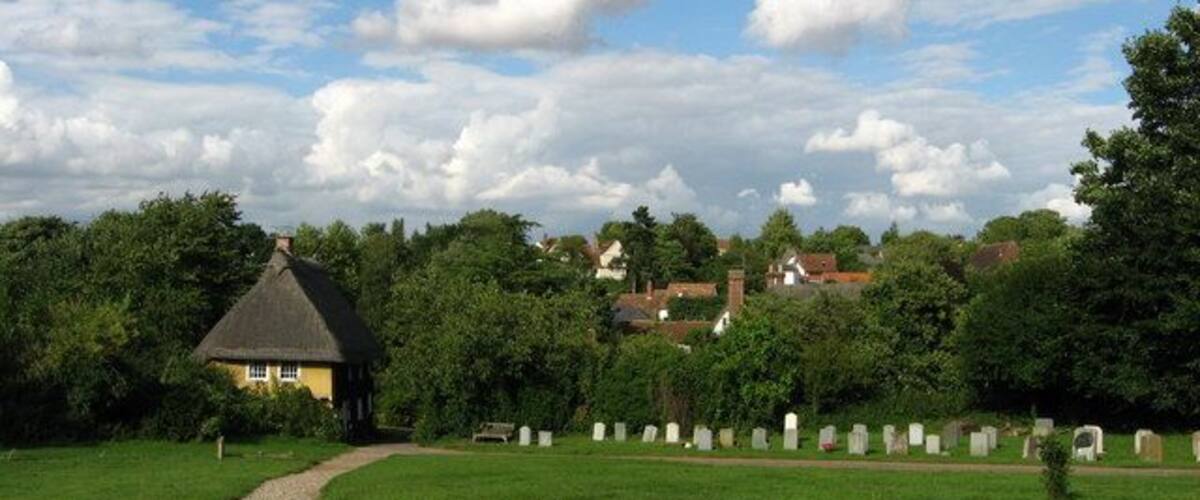 A rural cemetery with headstones, a thatched-roof house, and surrounding greenery.