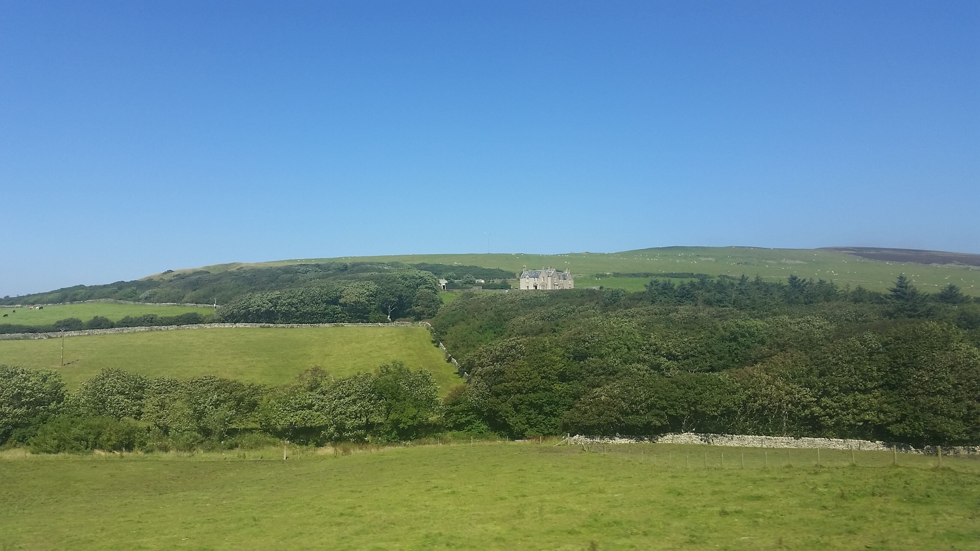 A green landscape with rolling hills, a stone wall, and a large building on a hill.