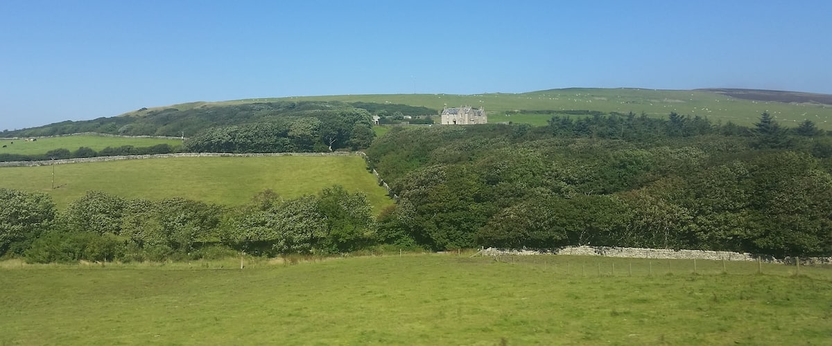 A green landscape with rolling hills, a stone wall, and a large building on a hill.