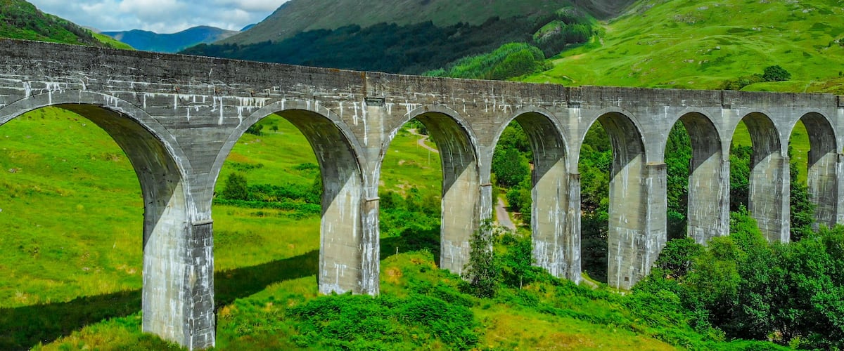 A stone viaduct with multiple arches over a lush green valley, with mountains in the background.