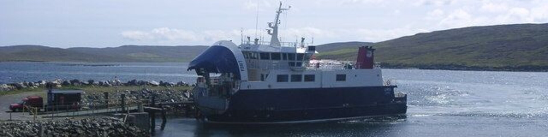 Un ferry atracado en un muelle, con un césped al frente y colinas en el fondo.