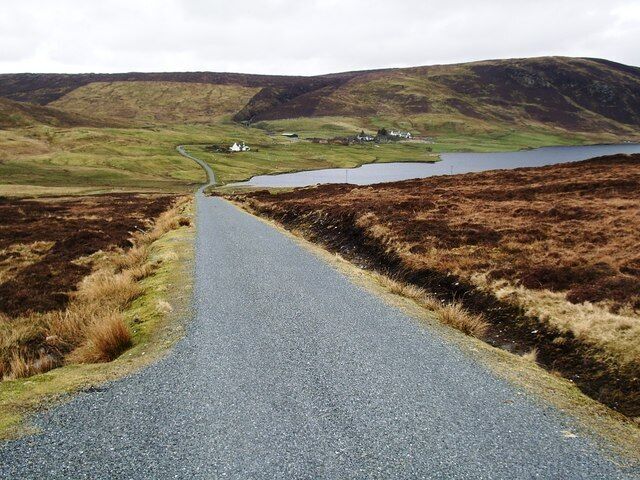 A narrow road winding through a hilly landscape with a lake and a small settlement in the distance.