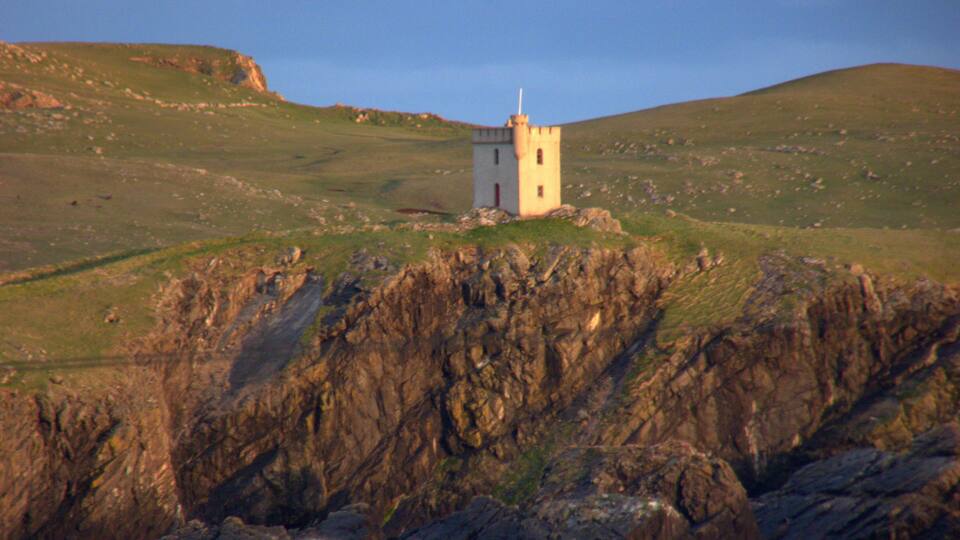 A small white tower on a grassy hill with rocky cliffs in the foreground.
