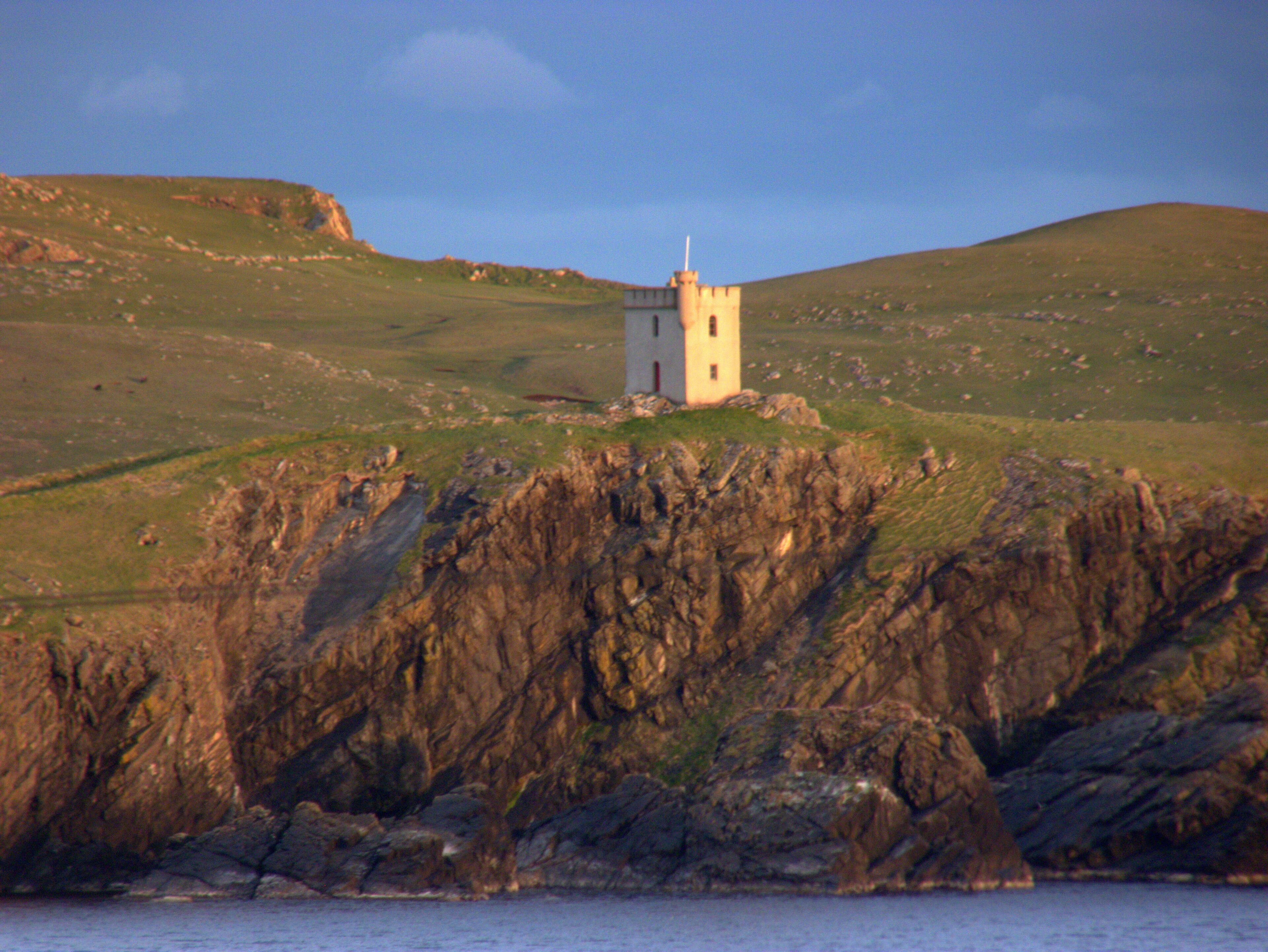 A small white tower on a grassy hill with rocky cliffs in the foreground.