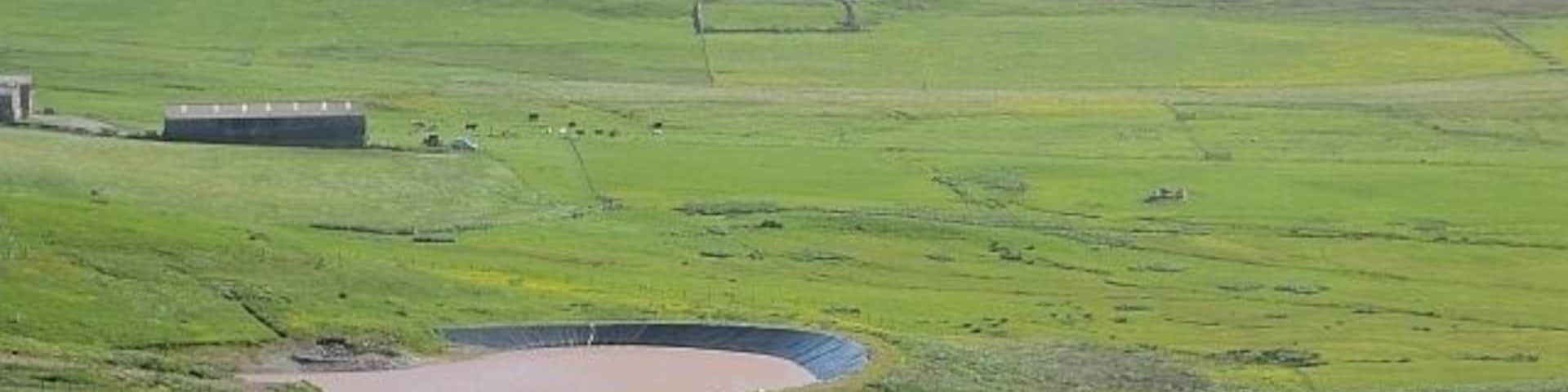 Un paisaje verde con una estructura circular, un edificio y un área cercada.