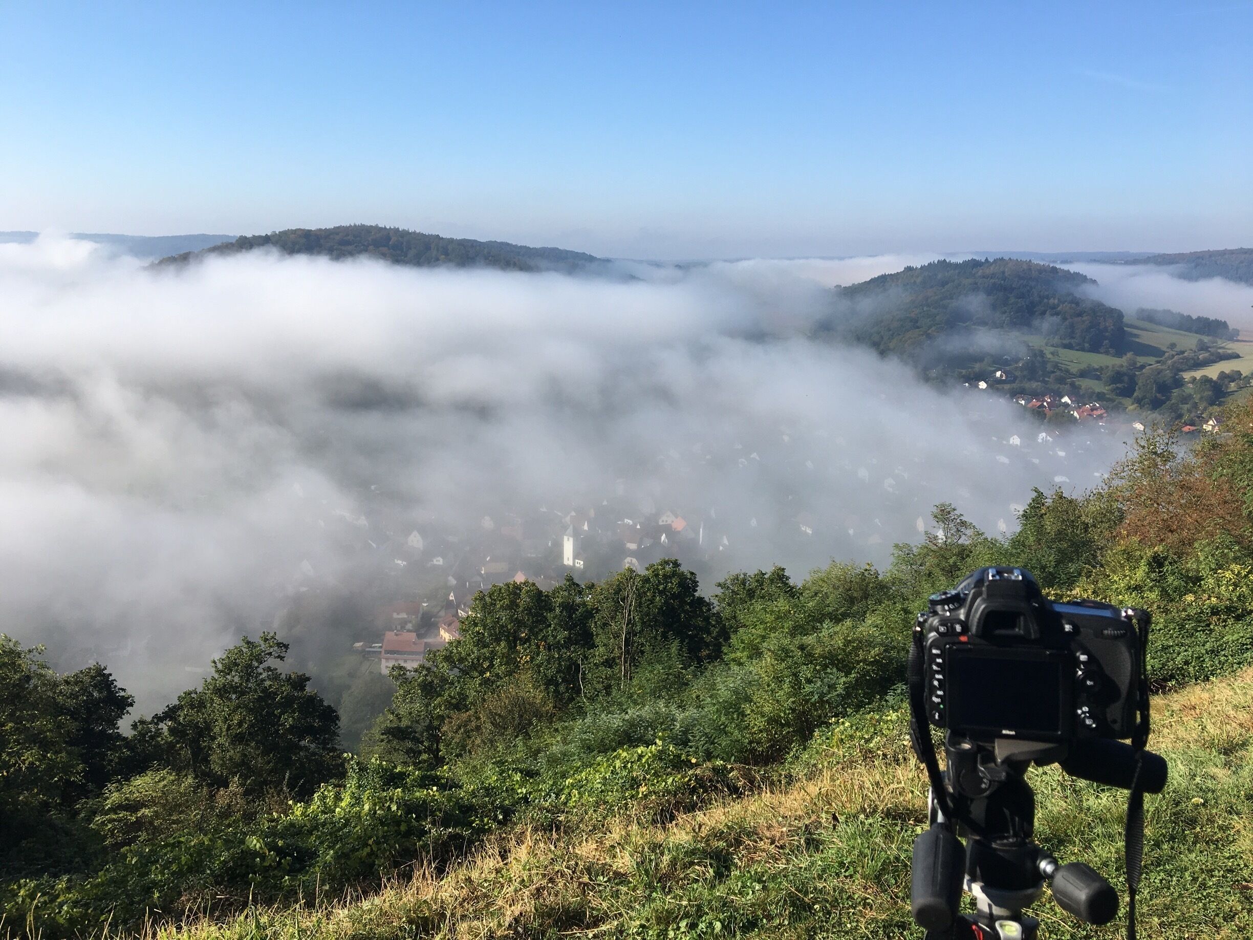 A camera on a tripod overlooking a valley with a town nestled among hills and enveloped in mist.