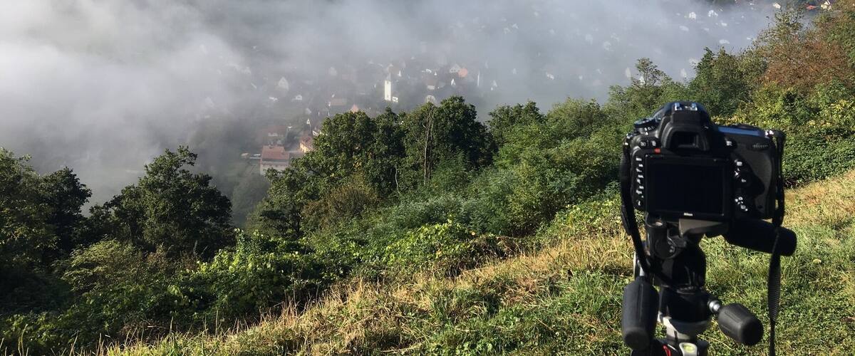 A camera on a tripod overlooking a valley with a town nestled among hills and enveloped in mist.