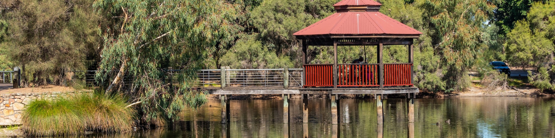 A red gazebo on a wooden pier over a calm lake, surrounded by dense trees and greenery.