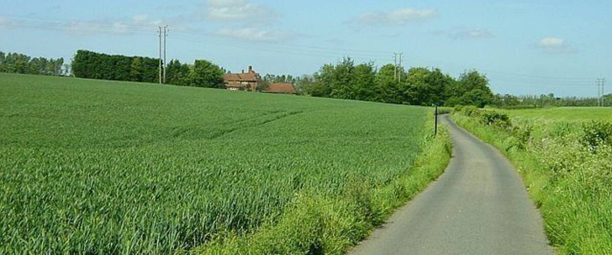 A narrow rural road flanked by green fields and a red-roofed house in the distance.