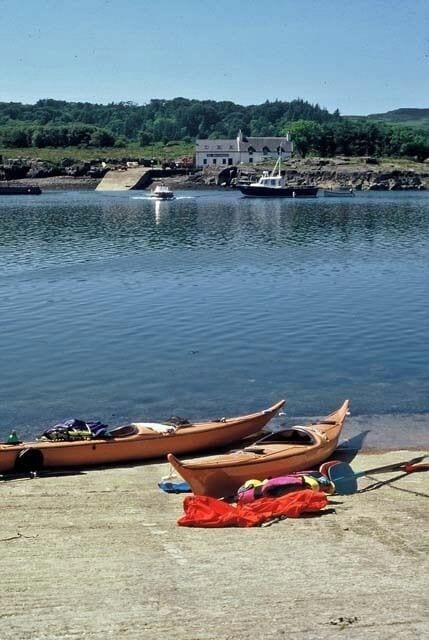 A calm harbor with two wooden boats docked, a small boat in the background, and a building on the shore.