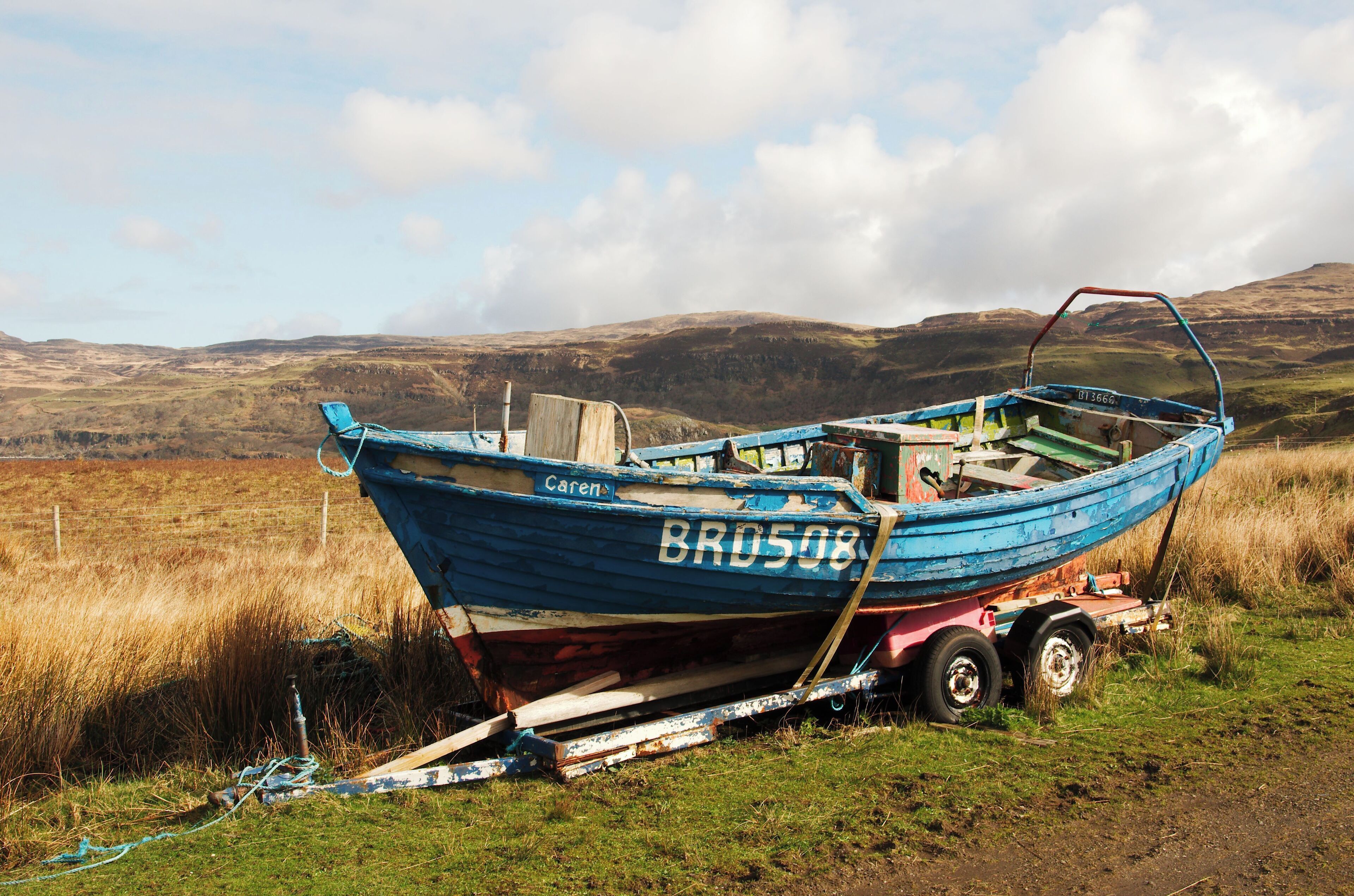 A blue boat named 'Bro508' with a trailer, parked on a grassy field with hills in the background.