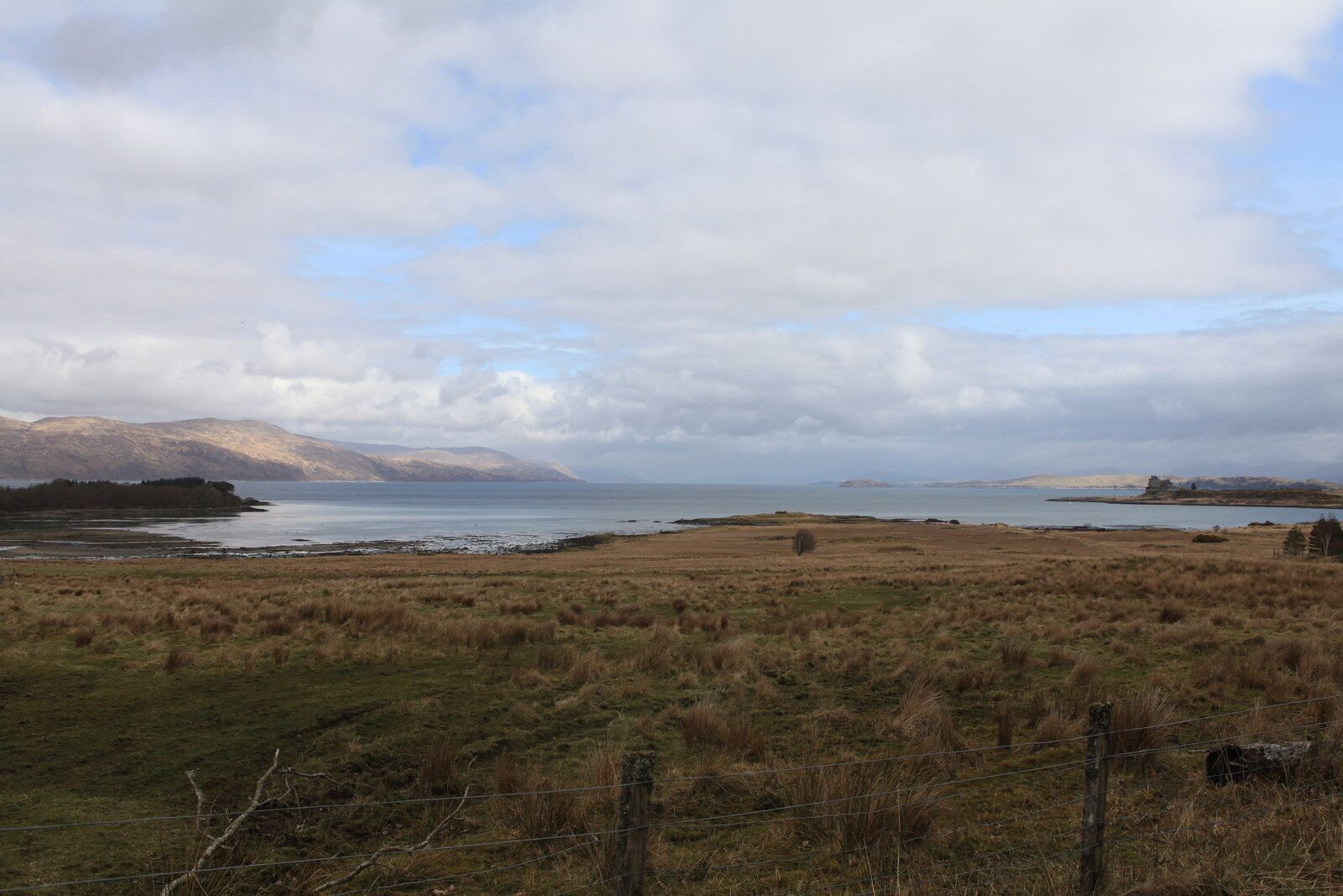 A coastal landscape with a grassy field, a body of water, and distant mountains under a cloudy sky.