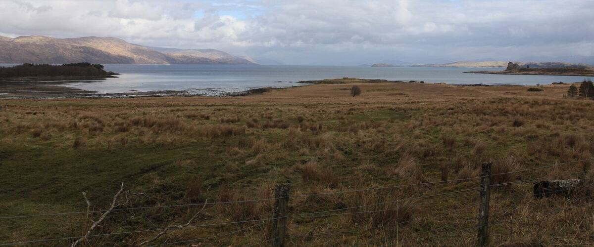 A coastal landscape with a grassy field, a body of water, and distant mountains under a cloudy sky.