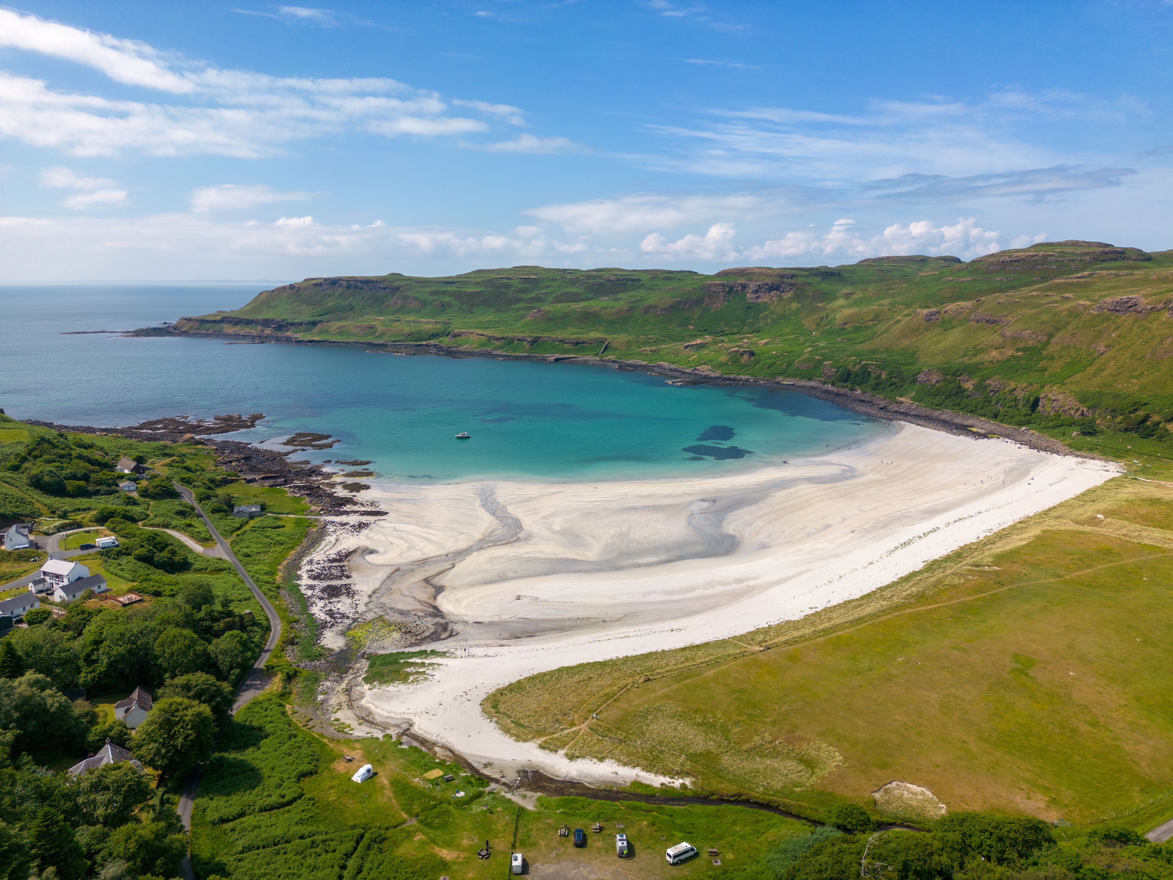 A coastal landscape with a sandy beach, green hills, and a clear blue sea.