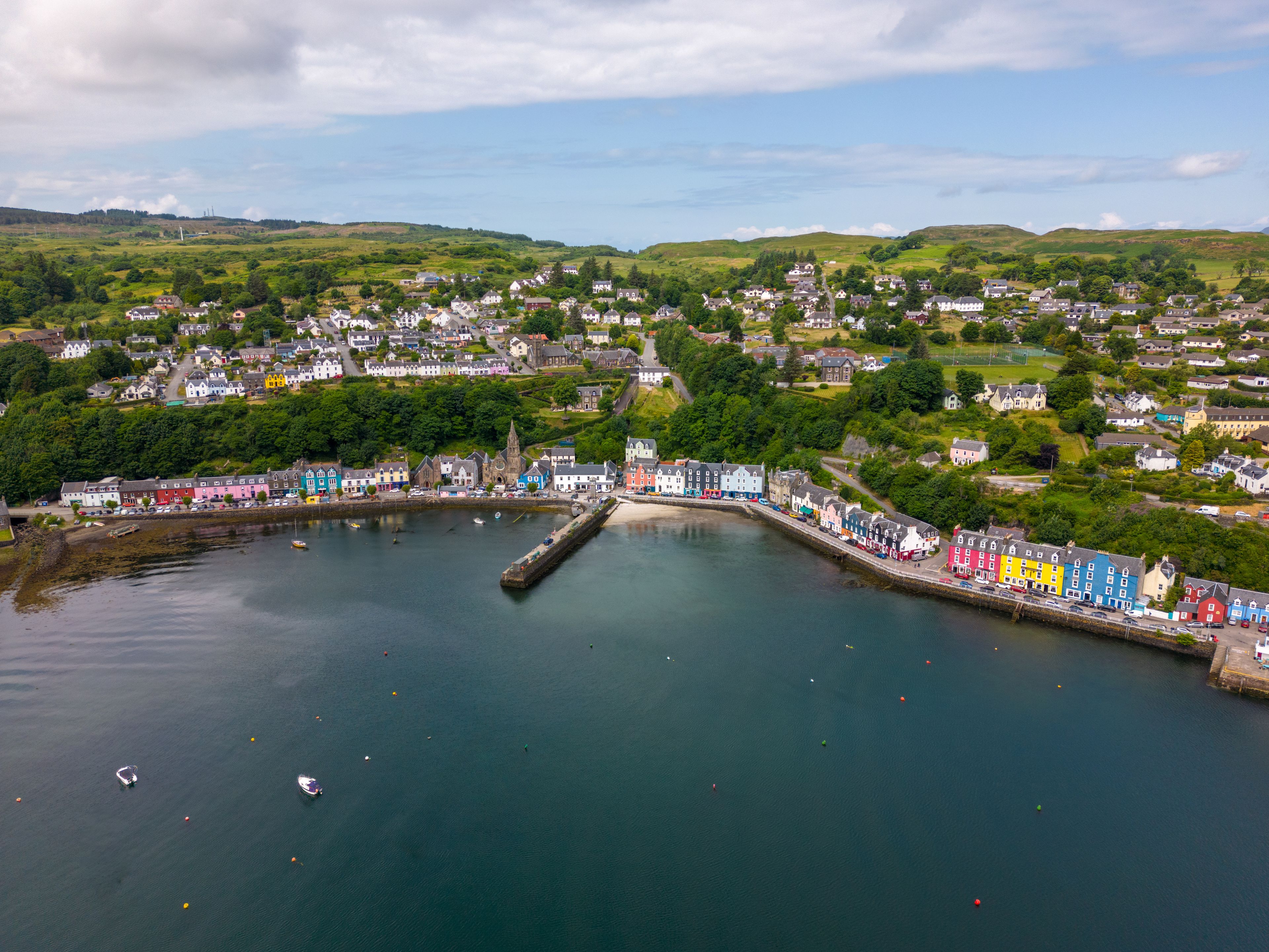 A coastal town with colorful buildings along the waterfront, a pier, and rolling hills in the background.