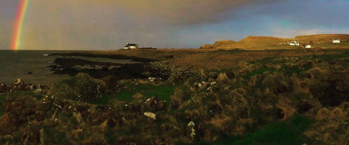 A coastal landscape with a rainbow, rocky terrain, and distant white buildings.