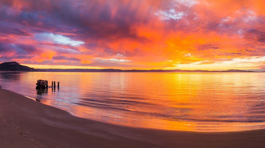 Una puesta de sol sobre un mar tranquilo con una playa arenosa y pilotes de madera en el agua.