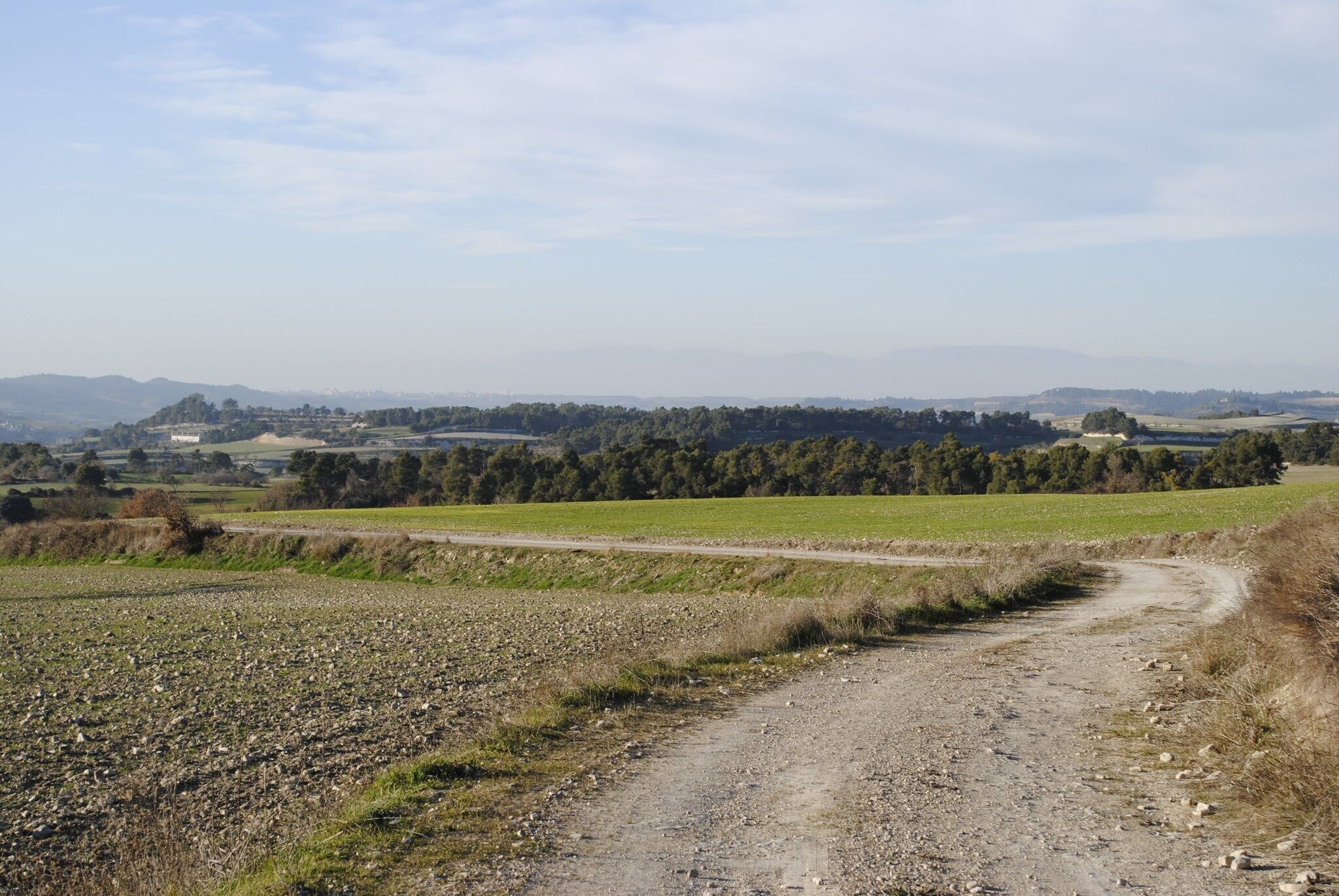 A rural dirt road winding through fields with distant hills and scattered buildings.