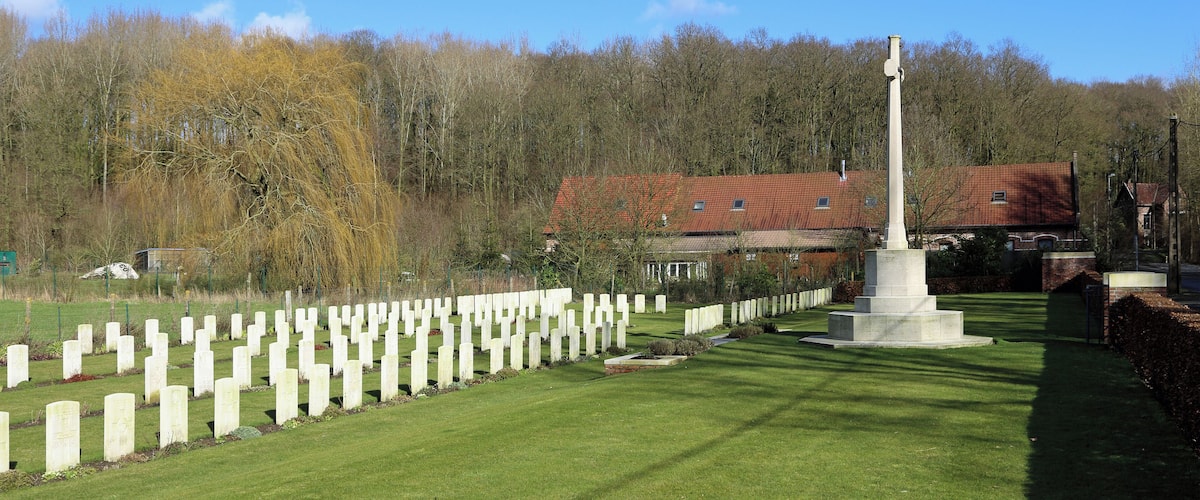 A cemetery with white headstones, a tall monument, and a red-roofed building in the background.