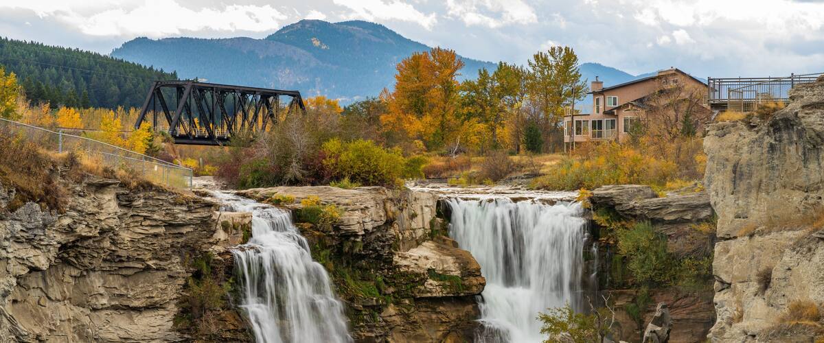 A waterfall cascading over rocky terrain with a bridge and buildings in the background.