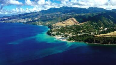 A coastal landscape with a clear blue sea, green hills, and a small town by the water.