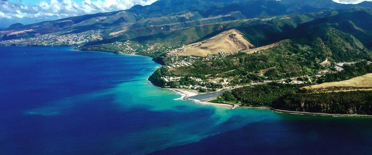 A coastal landscape with a clear blue sea, green hills, and a small town by the water.