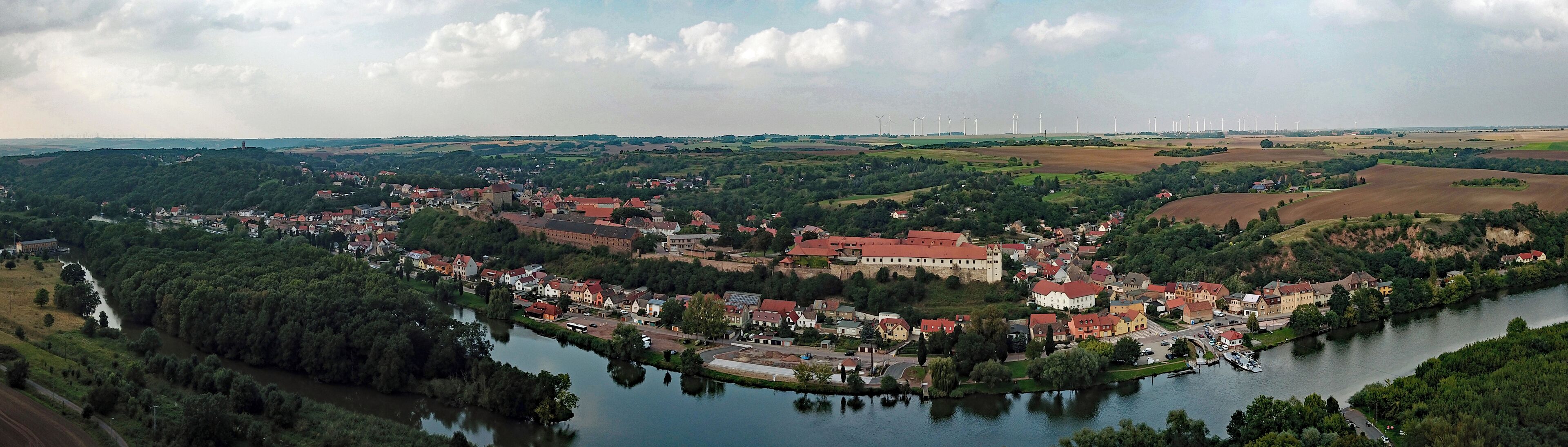 A town by a river with wind turbines in the distance.