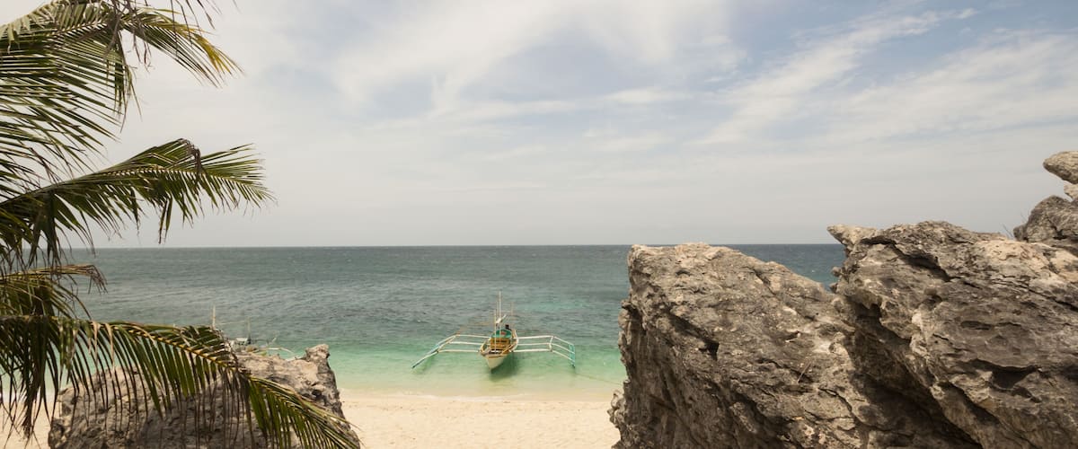 A beach with a boat anchored near rocky formations and palm trees.