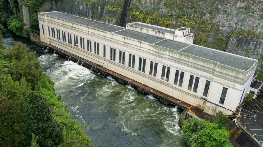Een grote witte dam met meerdere waterpoorten, omringd door weelderige begroeiing en een steile klif.