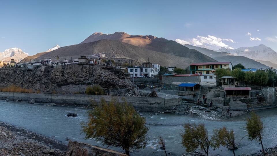 A river flowing through a mountainous village with buildings and a bridge.