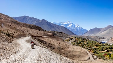 Un paisaje montañoso y agreste con un camino de tierra, una persona en motocicleta y un río.
