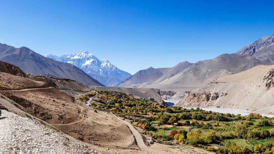 A rugged mountain landscape with a dirt road, a person on a motorcycle, and a river.