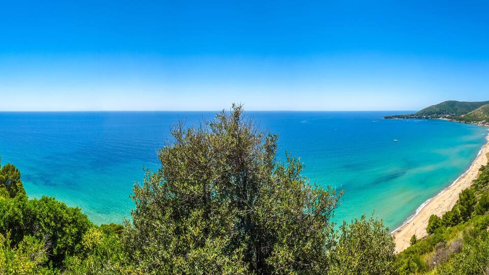 A coastal landscape with a clear blue sea, a sandy beach, and green hills.