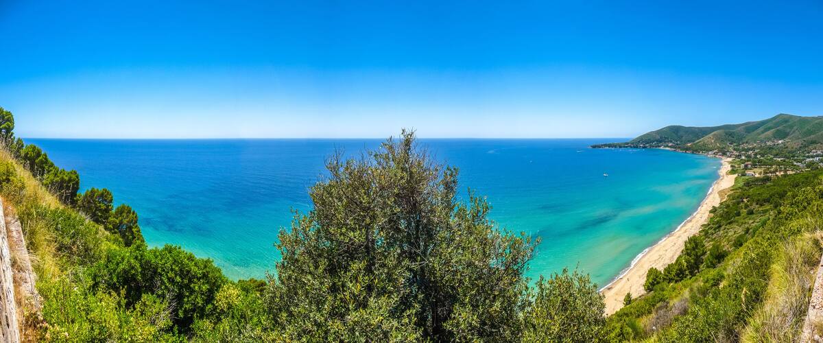 A coastal landscape with a clear blue sea, a sandy beach, and green hills.