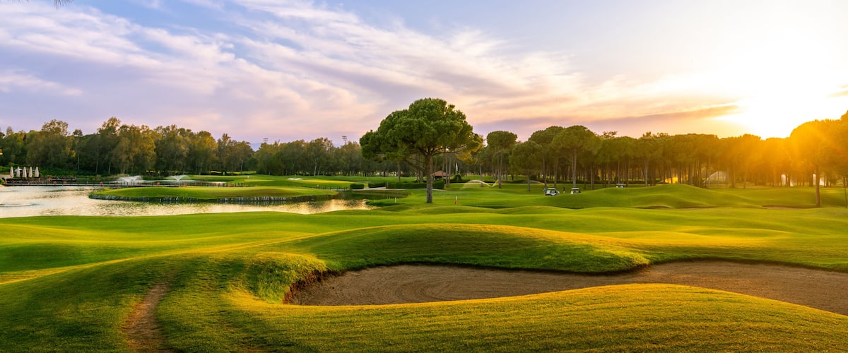 Golf course at sunset with beautiful sky and sand trap. Scenic panoramic view of golf fairway with bunker. Golf field with pines