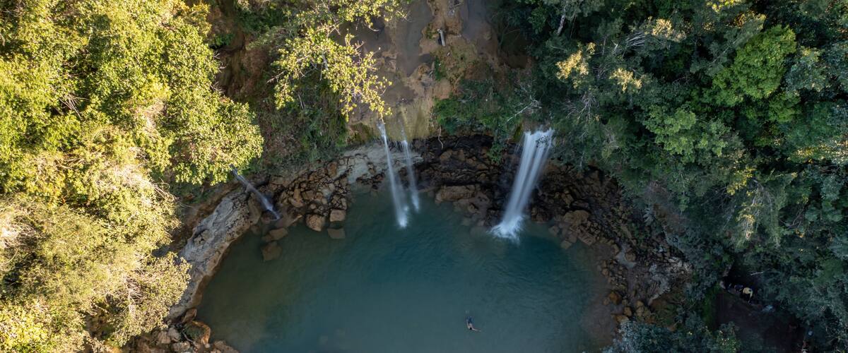An aerial view of a natural waterfall cascading into a circular pond surrounded by dense forest.