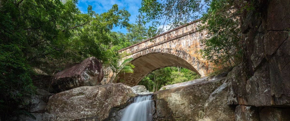 A stone bridge over a waterfall surrounded by large rocks and lush greenery.
