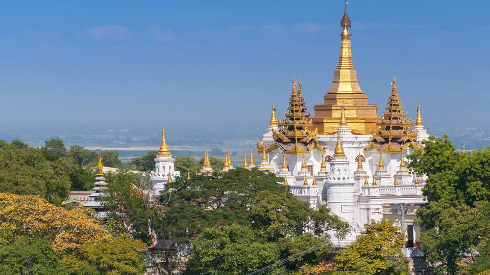 A large white temple complex with golden spires and multiple pagodas surrounded by lush green trees.
