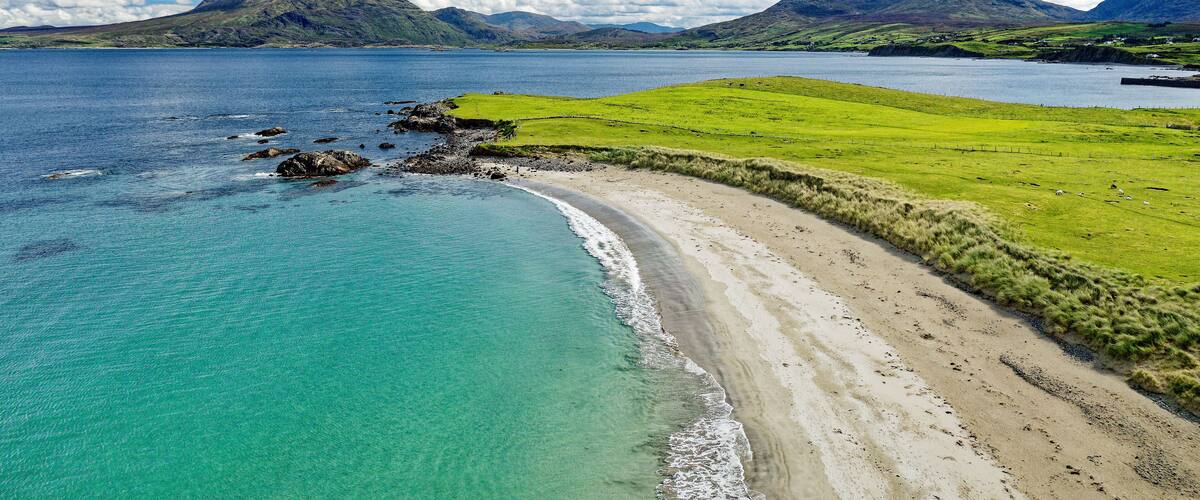 A coastal landscape with a clear blue sea, a sandy beach, and rolling hills in the background.