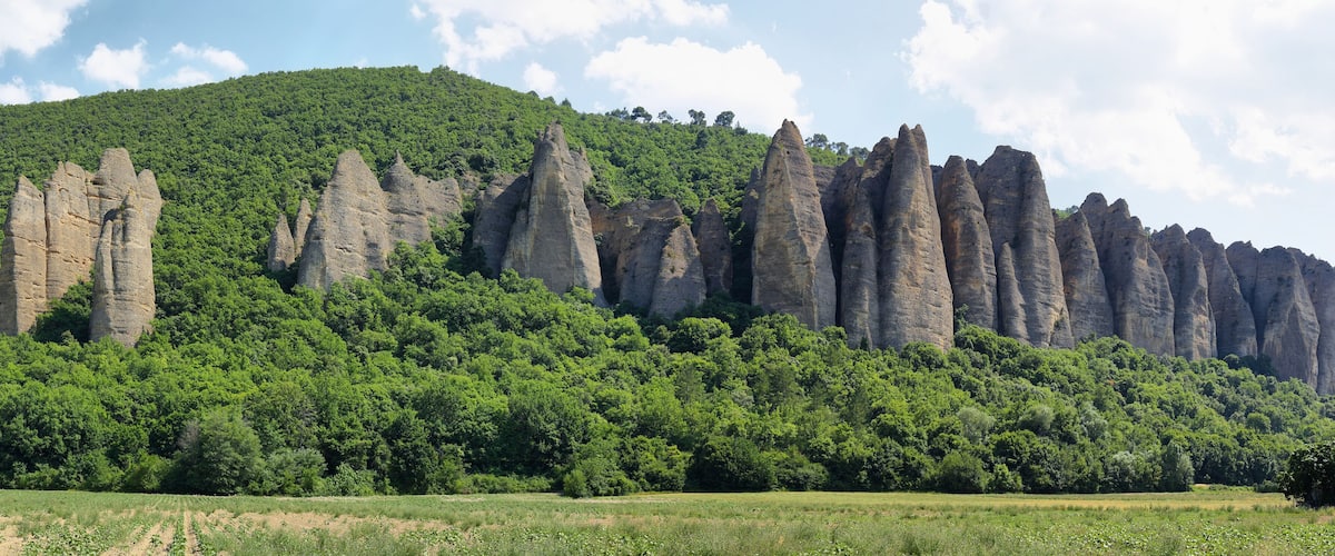A landscape with tall, narrow rock formations and a lush green hill in the background.