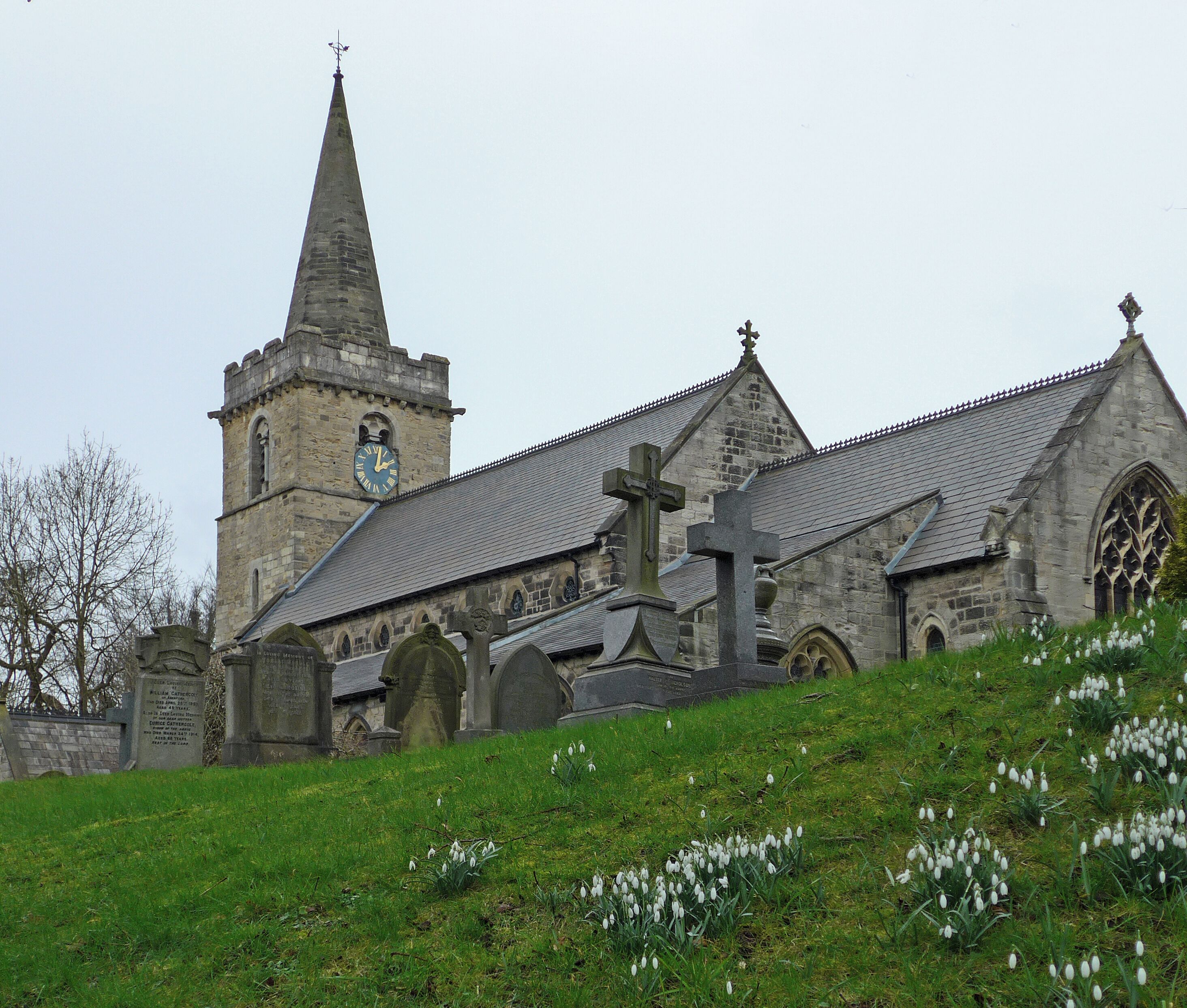 A church with a tall steeple and a clock, surrounded by gravestones and snowdrops.
