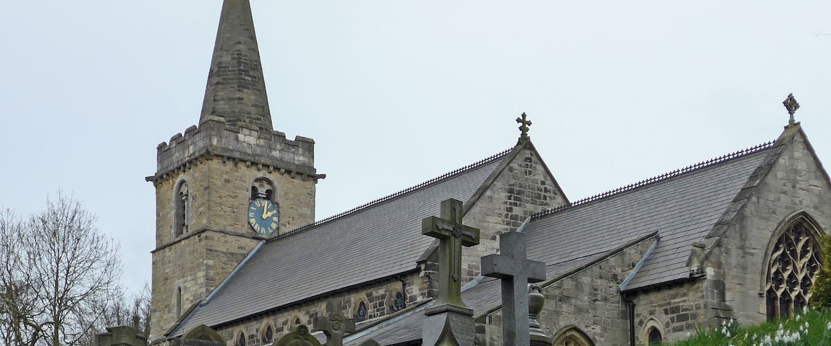 A church with a tall steeple and a clock, surrounded by gravestones and snowdrops.