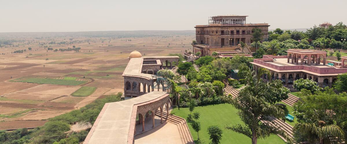 A historical building complex with arched structures and a large dome, surrounded by lush greenery and terraced fields.