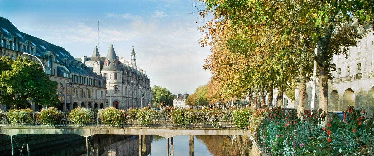 A canal with a bridge, a historic building with towers, and a tree-lined walkway with flowers.