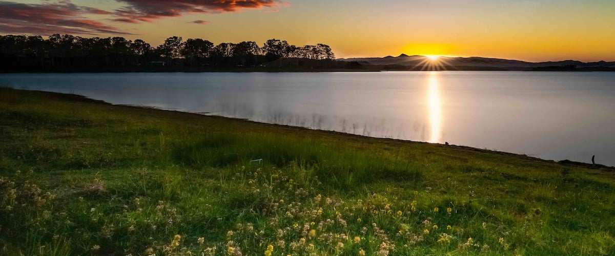 A sunset over a calm lake with grassy banks and a distant tree line.