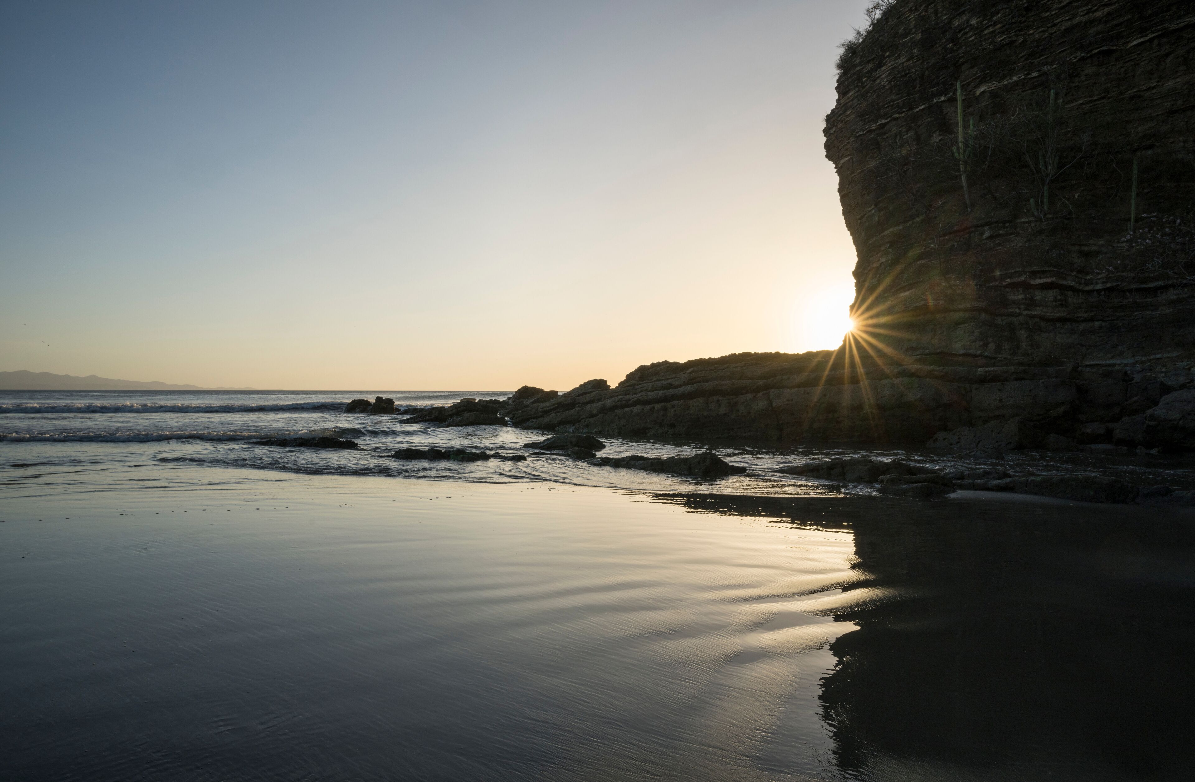 A beach with calm waters, a rocky cliff, and the sun setting on the horizon.